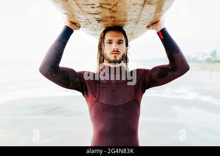 Porträt eines jungen Surfers mit langen Haaren und Bart in Neoprenanzug stehend Blick auf die Kamera am Strand mit dem Surfbrett über dem Kopf während Stockfoto