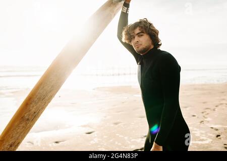 Seitenansicht eines lächelnden Surfers in Neoprenanzug, der während des Sonnenaufgangs im Hintergrund die Kamera mit dem Surfbrett am Strand betrachte Stockfoto
