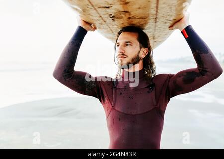 Porträt eines jungen Surfers mit langen Haaren und Bart in Neoprenanzug stehend mit Blick weg auf den Strand mit dem Surfbrett über dem Kopf während der Sonne Stockfoto