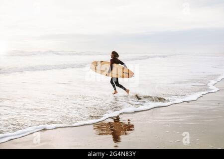 Seitenansicht des Surfermanns in Neoprenanzug, der mit Surfbrett zum Wasser läuft, um während des Sonnenaufgangs eine Welle am Strand zu fangen Stockfoto