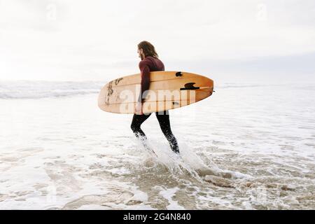 Seitenansicht des Surfermanns in Neoprenanzug, der mit Surfbrett zum Wasser läuft, um während des Sonnenaufgangs eine Welle am Strand zu fangen Stockfoto