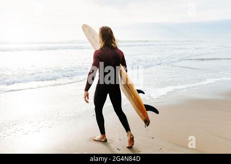 Seitenansicht eines anonymen Surfers, der in Neoprenanzug gekleidet ist und mit dem Surfbrett zum Wasser läuft, um während des Sonnenaufgangs eine Welle am Strand zu fangen Stockfoto