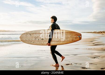 Seitenansicht des in Neoprenanzug gekleideten Surfers, der mit dem Surfbrett zum Wasser läuft, um während des Sonnenaufgangs eine Welle am Strand zu fangen Stockfoto