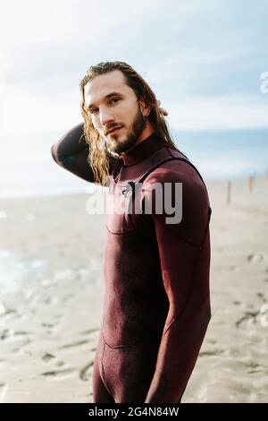 Seitenansicht eines jungen, hübschen Surfers mit langen Haaren in Neoprenanzug, der während des Sonnenaufgangs auf die Kamera am Strand blickt Stockfoto