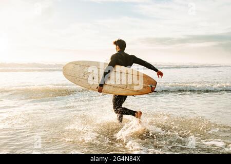 Seitenansicht des in Neoprenanzug gekleideten Surfers, der mit dem Surfbrett zum Wasser läuft, um während des Sonnenaufgangs eine Welle am Strand zu fangen Stockfoto