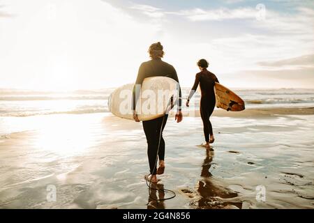 Rückansicht von nicht erkennbaren männlichen Surferfreunden in Neoprenanzügen, die mit Surfbrettern zum Wasser laufen, um während der Sonne eine Welle am Strand zu fangen Stockfoto