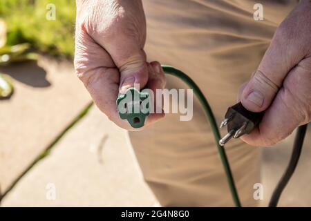 Nahaufnahme der Hände eines Mannes, die sich fertig machen, um das amerikanische Netzkabel mit drei Zapfen in das abgenutzte grüne Verlängerungskabel einzustecken Stockfoto