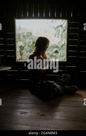 Ganzkörper-Seitenansicht einer ruhigen Frau, die in einer Holzhütte in der Nähe des Fensters in einem tropischen Land mit üppig grünen Pflanzen sitzt Stockfoto
