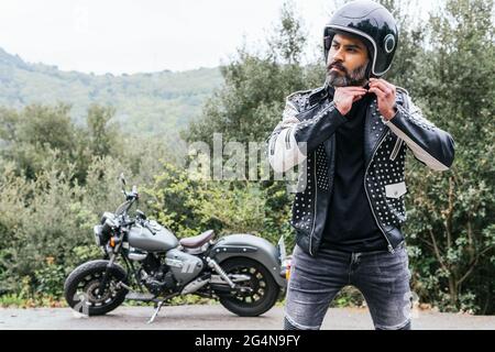 Selbstsicherer erwachsener bärtiger männlicher Biker in stilvoller Lederjacke, der den Schutzhelm verstellt und in die Natur hinwegschaut Stockfoto
