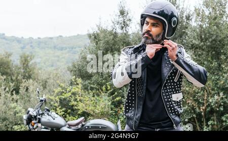 Selbstsicherer erwachsener bärtiger männlicher Biker in stilvoller Lederjacke, der den Schutzhelm verstellt und in die Natur hinwegschaut Stockfoto