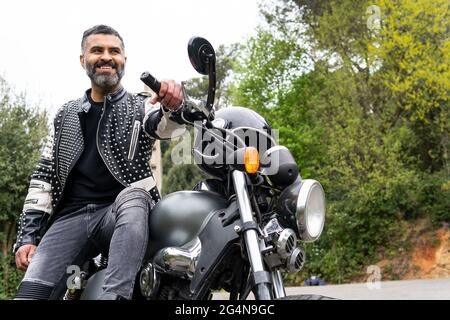 Von unten bärtiger, ethnischer Biker in schwarzer Lederjacke, der auf einem modernen Motorrad auf einer Asphaltstraße inmitten üppig grüner Bäume in den Bergen sitzt Stockfoto
