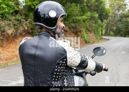 Rückansicht bärtiger ethnischer Biker in schwarzer Lederjacke und Helm auf einem modernen Motorrad auf Asphaltstraße inmitten üppig grüner Bäume, die in Moun wachsen Stockfoto