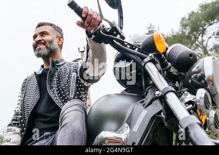 Von unten bärtiger, ethnischer Biker in schwarzer Lederjacke, der auf einem modernen Motorrad auf einer Asphaltstraße inmitten üppig grüner Bäume in den Bergen sitzt Stockfoto