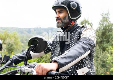 Bärtiger, ethnischer Biker in schwarzer Lederjacke und Helm, der auf einem modernen Motorrad auf einer Asphaltstraße inmitten üppig grüner Bäume im gebirgigen VA unterwegs ist Stockfoto