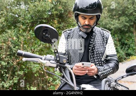 Männlicher Biker in trendiger Lederjacke mit Nieten und Schutzhelm Surfen Telefon, während auf Motorrad in der Nähe von üppigen grünen Wald geparkt sitzen Stockfoto