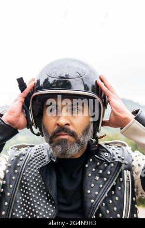 Selbstsicherer erwachsener bärtiger männlicher Biker in stilvoller Lederjacke, der den Schutzhelm verstellt und in die Natur hinwegschaut Stockfoto