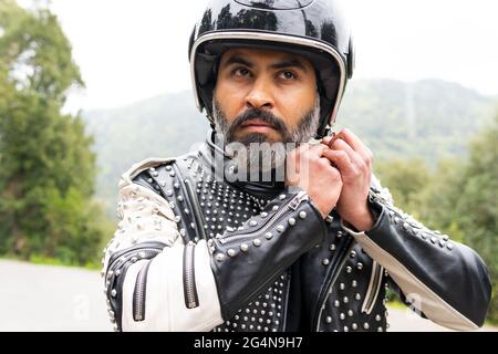 Selbstsicherer erwachsener bärtiger männlicher Biker in stilvoller Lederjacke, der den Schutzhelm verstellt und in die Natur hinwegschaut Stockfoto