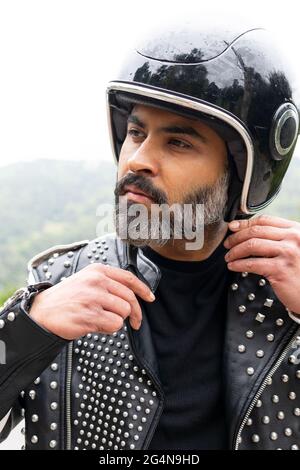 Selbstsicherer erwachsener bärtiger männlicher Biker in stilvoller Lederjacke, der den Schutzhelm verstellt und in die Natur hinwegschaut Stockfoto