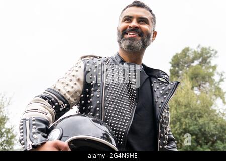Von unten brutal ethnischen männlichen Biker in stilvoller Lederjacke auf Asphalt Straße mit Helm in der Hand in der Nähe von üppigen grünen Bäumen wachsen in Mountainou Stockfoto