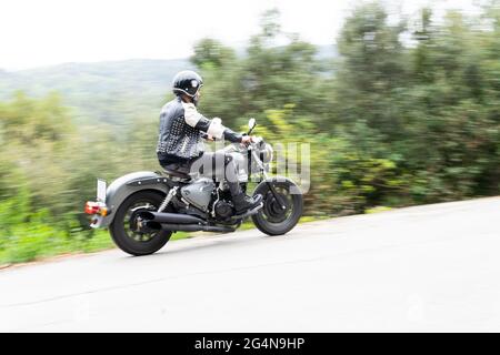 Ganzer Körper von bärtigen ethnischen männlichen Biker in schwarzer Lederjacke und Helm Reiten moderne Motorrad auf Asphaltstraße inmitten üppig grünen Bäumen in m wachsen Stockfoto