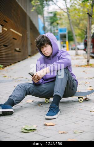 Ernsthafter männlicher Teenager in legerer Kleidung, der auf dem Skateboard sitzt und die Kamera anschaut, während er auf dem gepflasterten Bürgersteig in der Stadt Handy surft Stockfoto