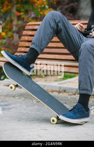 Nicht erkennbarer Mann in Sneakers balanciert auf Skateboard, während er auf einer Holzbank im Park mit Büschen während des Trainings in der Stadt sitzt Stockfoto