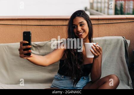 Glückliche junge hispanische Frau mit einem Becher heißen Getränks, lächelnd und Selfie, während sie auf dem Sofa auf dem Balkon sitzt Stockfoto