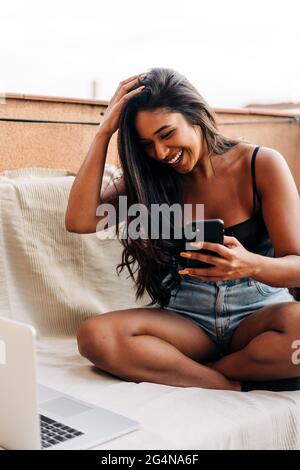 Optimistische junge hispanische Frau, die Haare berührt und lacht, während sie auf der Couch neben dem Laptop sitzt und auf dem Balkon auf dem Smartphone surft Stockfoto