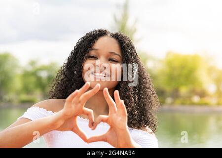 Lächelnde junge schwarze Dame in Sommerkleidung in der Nähe von See und grünen Bäumen im Park, die auf die Kamera schaut, während sie an sonnigen Tagen mit den Händen eine Herzbewegung macht Stockfoto