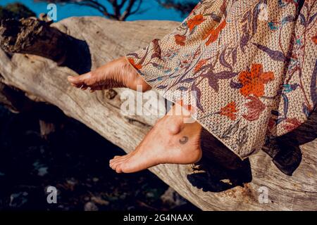 Unkenntlich Ernte Frau im Sommerkleid und mit Tätowierung auf Bein sitzt auf Baumstamm im Wald Stockfoto