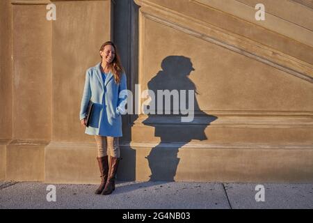 Ganzer Körper von glücklichem erwachsenen Weibchen in blauem Mantel, der in der Nähe einer Steinmauer mit einem Ordner in den Händen in der Stadtstraße an sonnigen Tagen steht Stockfoto