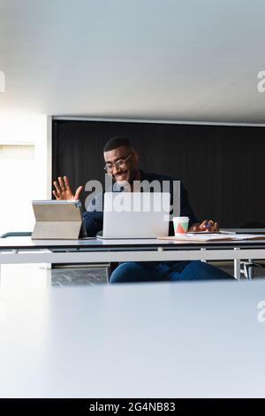 Positiver afroamerikanischer Erwachsener, männlich in formeller Kleidung und Brillen, die am Tisch sitzen, während er einen Videoanruf auf dem Tablet in der Nähe des Netbooks führt und Wi dokumentiert Stockfoto