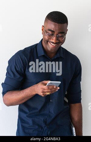 Afroamerikanischer erwachsener Mann in stilvollem Outfit und Brille, die mit einem Netbook in den Händen steht, während das Mobiltelefon auf weißem Hintergrund verwendet wird Stockfoto