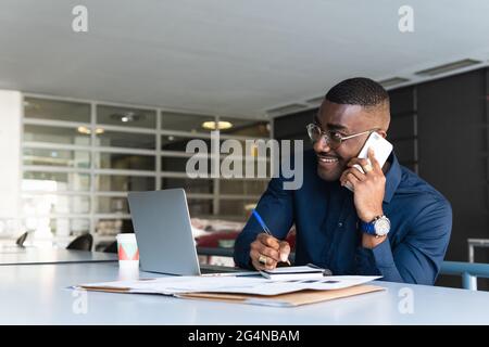 Lächelnder afroamerikanischer Mann in Brillen und formeller Kleidung am Tisch, der sich mit dem Handy unterhält und sich in einem Notizblock neben Netbook und Notizen machte Stockfoto