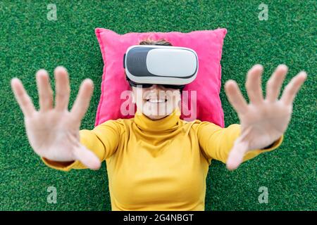 Von oben lächelnde Frau in legerer Kleidung, die auf einem Kissen auf grünem Teppich liegt, während sie virtuelle Realität im VR-Headset mit erhobenen Händen bei c erlebt Stockfoto