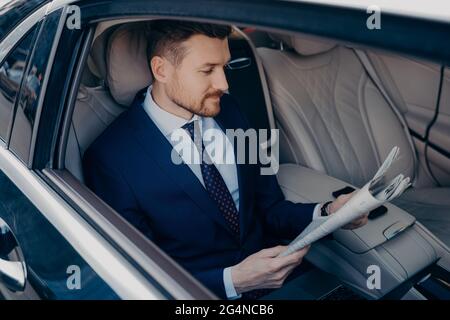 Durchdachter Manager in formellem Anzug, der Zeitung auf dem Rücksitz der Limousine liest Stockfoto