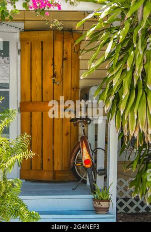 Eingang und Veranda zum Key West Haus mit rustikalem Hurrikan-Verschluss an der Tür und verrosteten Fahrrad, umgeben von tropischem Grün geparkt Stockfoto