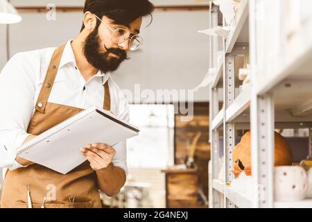 Junger attraktiver Geschäftsmann ein Töpfer mit Bart und Schnurrbart arbeitet in seiner Werkstatt. Speichert Aufzeichnungen und transkribiert sie in einem Notizbuch, das überprüft wird Stockfoto