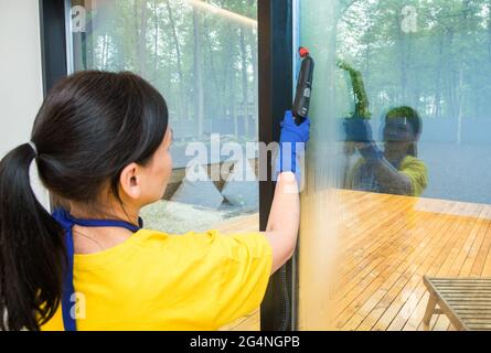 Professioneller Reinigungsservice. Frau in Uniform und Handschuhen Schwamm wäscht Panoramafenster in der Hütte. Dampfwischen und Schaber waschen Stockfoto