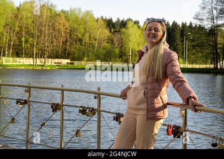 Die junge kaukasische, dicke Dame steht lässig auf einer Brücke im Park und schaut in die Kamera. Fröhliche, fröhliche Frau, die die Wochenenden und das gute Wetter genießt Stockfoto