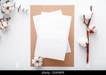 Weißes, unbeschriftete Karte-Mockup-Notizpapier mit trockenen blühenden Baumwollblumen auf flachem Lay. Modernes Desktop-Modell für Grußkarten. Eleganter Arbeitsbereich mit Stockfoto