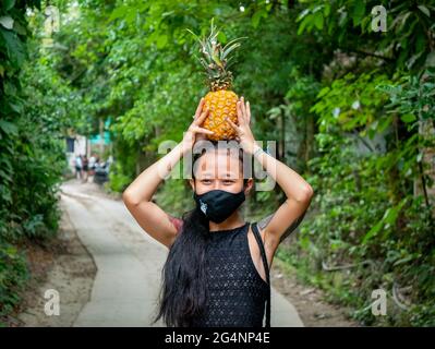 Eine asiatische Frau mit einer schwarzen Maske im Mund hält eine Ananas auf ihrem Kopf Stockfoto