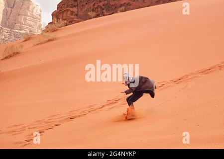 Junger Mann, der Sanddüne surft, trägt bisht - einen traditionellen Beduinenmantel. Sandsurfing ist eine der Attraktionen in der Wadi Rum Wüste Stockfoto