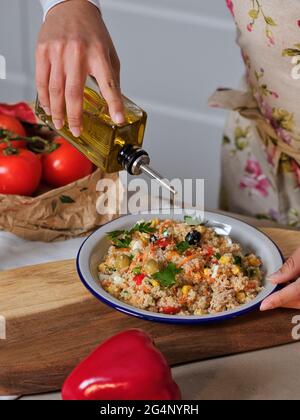 Die Hände der Latina-Frau tröpfeln Olivenöl auf den Teller mit Quinoa-Salat in der heimischen Küche, Nahaufnahme von rotem Paprika und Papiertüte im Hintergrund mit Tomaten Stockfoto
