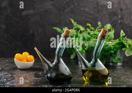 Zwei Glas-Olivenölausgiesser mit Korken auf dunklem Hintergrund. Gelbe Tomaten in einer Schüssel und einige Pflanzen in einem Becken dahinter. Stockfoto