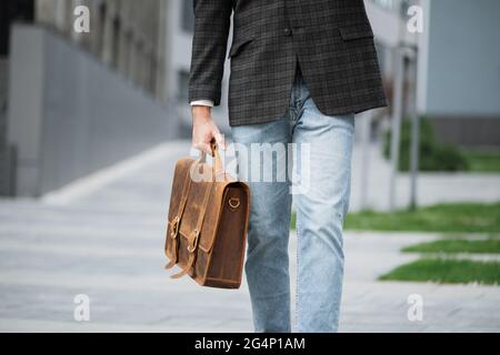 Nahaufnahme eines jungen Mannes in blauer Jeans und formeller Jacke, der einen braunen Lederkoffer trägt, während er in der Nähe des Bürogebäudes läuft. Konzept von erfolgreichen Menschen, Wirtschaft und Karriere. Stockfoto
