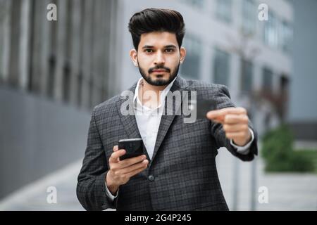 Erfolgreicher arabischer Geschäftsmann, der im Freien steht und ein modernes Smartphone mit Bankkarte hält. Bärtiger Mann in formeller Kleidung, der online Einkäufe tätigt. Stockfoto