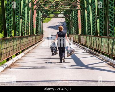 Eine kaukasische Frau mittleren Alters mit roten Haaren, schwarzen Kleidern und Handschuhen trägt Plastik und einen Müllbeutel über eine Metallbrücke, um eine Freiwillige aufzuräumen. Stockfoto