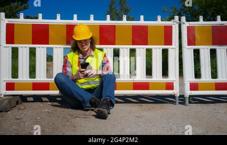 Männlicher Arbeiter in Hardhut und hoher Visusjacke mit Smartphone. Stockfoto