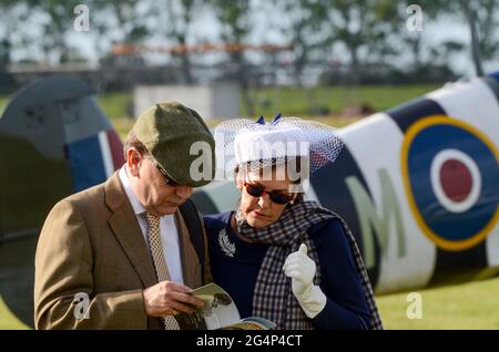 Besucher des Goodwood Revival in Vintage-Kleidung. Männliches und weibliches Paar in zeitlichem Kostüm, mit einem Spitfire-Flugzeug. Nostalgie-Ereignis. Öffentliche Eintauchen Stockfoto